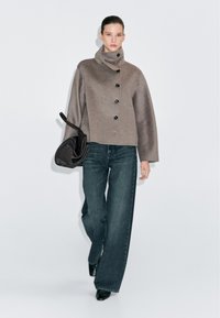 Giacca corta marrone con colletto alto e bottoni neri, abbinata a jeans larghi scuri. La modella tiene una borsa nera. Sfondo semplice.