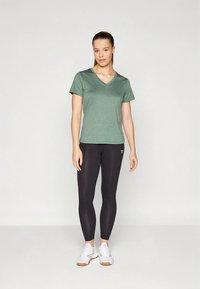 Maglietta verde a scollo a V in tessuto leggero e strutturato, abbinata a leggings neri e scarpe da ginnastica bianche. Design semplice con maniche corte.