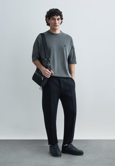 Šedé tričko s krátkým rukávem s jemným logem, kombinované s černými kalhotami s plisováním a černými teniskami. Součástí je malá černá kabelka přes rameno.