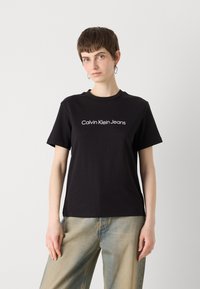 Čierne bavlnené tričko s krátkymi rukávmi a okrúhlym výstrihom; na prednej strane má biele logo "Calvin Klein Jeans". V kombinácii so svetlými džínsami.