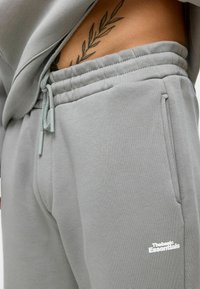 Grijze katoenen joggers met elastische tailleband, zijzakken en trekkoord. Kenmerkt zich door een wit logo op de onderste linkerpijp. Zachte textuur.