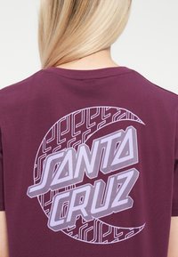 Burgunderfarget bomulls-t-skjorte med stor grafisk design i lyse lilla farge, med teksten "SANTA CRUZ" og geometriske mønstre bak.