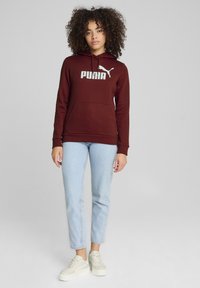 Felpa color bordeaux con logo bianco "PUMA", tasca a marsupio e cappuccio con cordino. Abbinata a jeans azzurri chiari e sneakers color crema.