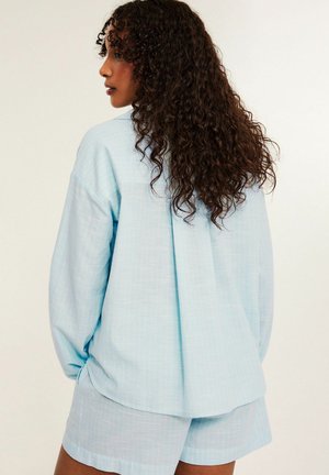 Frau mit langen lockigen Haaren, die ein locker sitzendes hellblaues Hemd und passende Shorts trägt, von hinten vor einfachem Hintergrund gesehen.