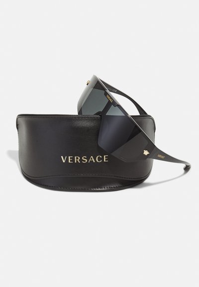 Versace sunglasses with black frames and dark lenses resting on a black leather case embossed with gold "Versace" logo.