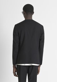 GIacca nera su misura, forma strutturata, texture liscia, spacchetto singolo sul retro, quattro bottoni sulla manica, indossata sopra una camicia bianca.