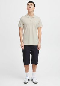 Beige polo skjorte med krage, korte ermer og en liten logo på brystet. Kombinert med mørke shorts og hvite sportsokker og sko.