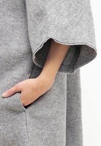 Main partiellement insérée dans la poche d'un manteau en laine gris à larges manches.