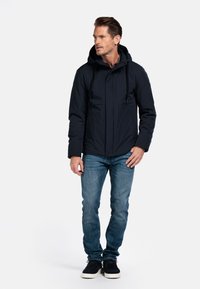 Marineblauw hoodie jack met een gladde textuur, voorzien van voorzakken met rits en verstelbare koorden, gecombineerd met blauwe jeans en donkere schoenen.