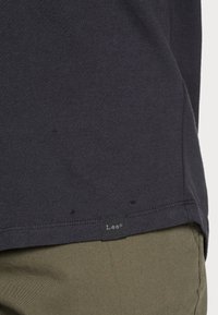 Gros plan sur un ourlet de chemise gris foncé avec une petite étiquette noire portant la mention "Lee" au-dessus d'un pantalon vert olive.
