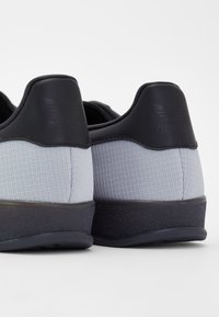 Gray sneakers featuring a textured grid pattern, black accents, and a rubber outsole. Soft material at the heel with embossed branding.