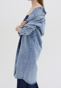 Vaalean sininen farkkukangas ylipitkä takki rentoa istuvuutta, napitettava sulkeminen ja käärityt hihat. Sileä pinta ja kaareva helma.