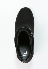 Bottine noire en daim doux, avec une forme élégante, des coutures discrètes et une bande décorative avec des accents métalliques.