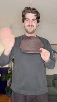 Pruun snapback-müts ümaralt reljeefse logoga. Pehme materjal ja struktureeritud kuju. Kandja on halli pika varrukaga särgi riides.