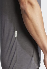 Szürke, rövid ujjú póló sima textúrával, oldalsó címkével és kontrasztos varrási részletekkel az alján és az ujj szélén.