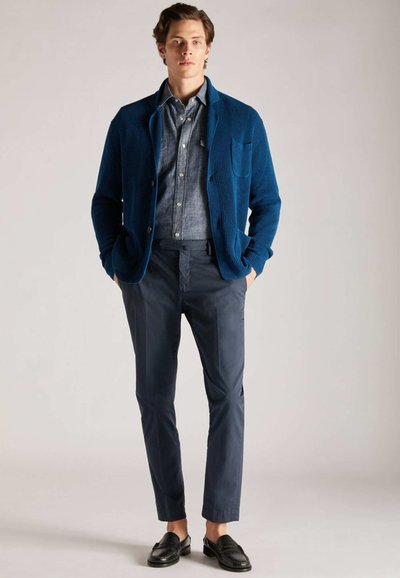 Blauwe gebreide cardigan over een spijkerhemd, gecombineerd met marineblauwe broek en zwarte instappers; heeft knoopdetails en een zak op de cardigan.