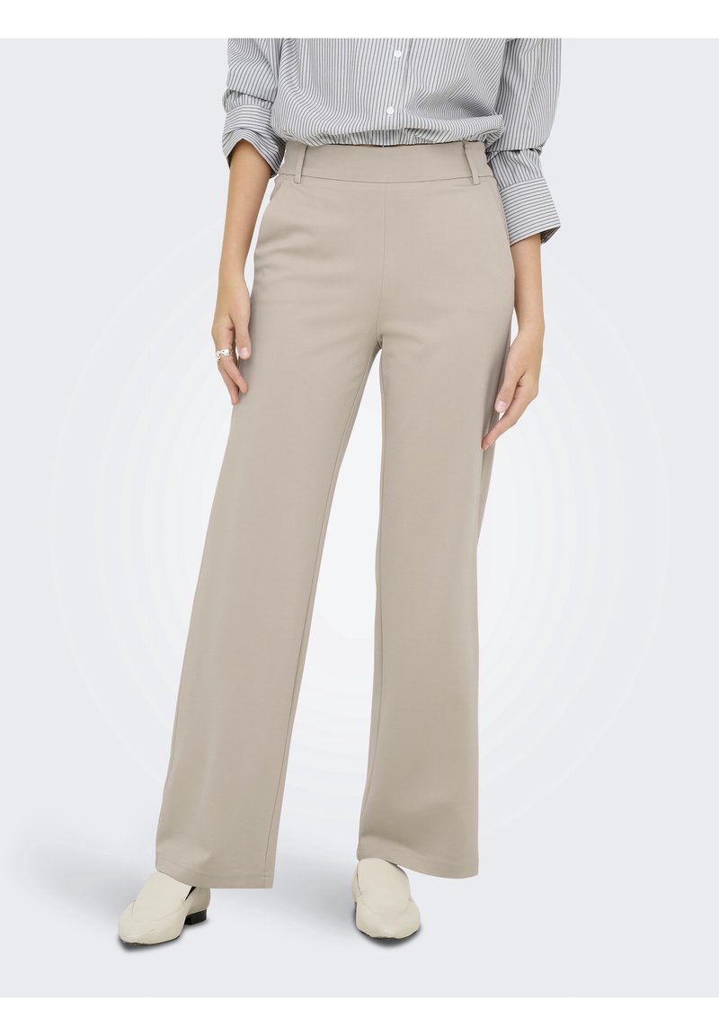 Femme portant un pantalon beige à jambes larges, des chaussures blanches sans lacets et une chemise rayée grise boutonnée avec manches retroussées.
