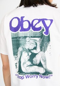 Hvit kortermet t-skjorte med et lilla og grønt grafisk design som har teksten "OBEY" og "Stop Worry Now!" over en illustrasjon av en sittende figur.