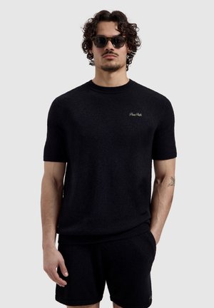 Man die een zwart gestructureerd shirt met korte mouwen en bijpassende short draagt, donkere zonnebril, staand met één hand in de zak tegen een eenvoudige achtergrond.