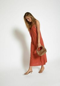 Jeune femme dans une robe sans manches orange tenant un sac à main en osier marron, portant des sandales à talons sangle beiges, se tenant devant un fond blanc uni.