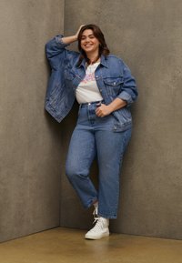 Femme souriante en veste en denim et jeans, adossée à un mur gris, portant des baskets blanches et un t-shirt graphique blanc.