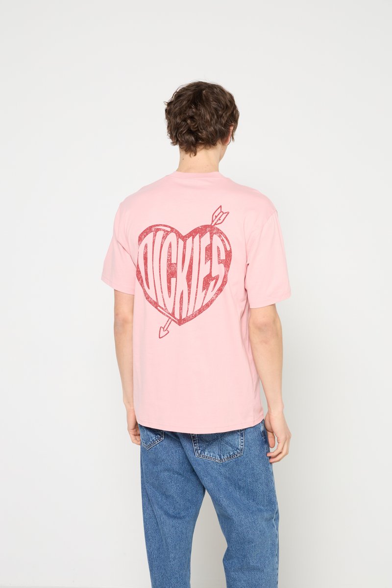 Personne portant un t-shirt rose avec un graphique d'un cœur rouge et une flèche contenant le mot "Dickies" au dos, associé à un jean bleu.