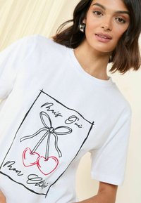 T-shirt en coton blanc avec un design graphique noir et rouge de cerises avec un nœud, texte "Mon Chéri" et contour minimaliste.