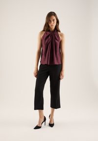 Top halter in raso color prugna con design anteriore plissettato, abbinato a pantaloni neri corti e décolleté nere con punta a punta su uno sfondo chiaro.