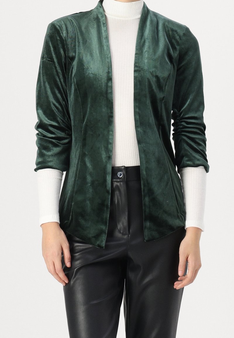 Grön sammetsp blazer med sjalkrage, öppen fram och långa ärmar. Kombinerad med en vit ribbad polokrage och svarta läderbyxor.