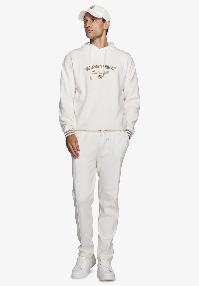 Hvid hoodie med "VARSITY TEAM" broderet i brunt, parret med matchende hvide bukser og hvide sneakers. Har snoretræk og ribbede manchetter.