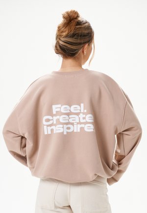 Frau mit blondem Haar im Dutt, die einen beigen Pullover mit weißem Schriftzug „Feel. Create Inspire.“ auf dem Rücken trägt, kombiniert mit hellfarbigen Hosen.