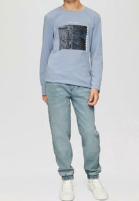 Light blue long-sleeve shirt with a graphic print featuring buildings; paired with light denim jeans and white sneakers with textured accents.
