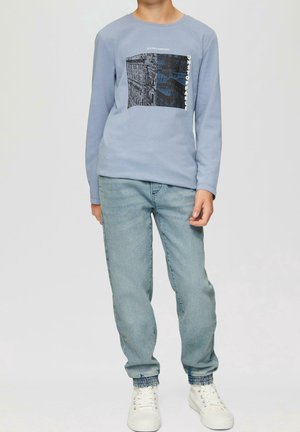 Camisa de manga larga de color azul claro con un estampado gráfico que presenta edificios; combinada con jeans de mezclilla claros y zapatillas blancas con acentos texturizados.