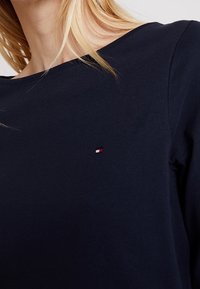 Námořnická modrá bavlněná košile s kulatým výstřihem, na levé části hrudi s malým červeným, bílým a modrým logem. Dlouhé rukávy.