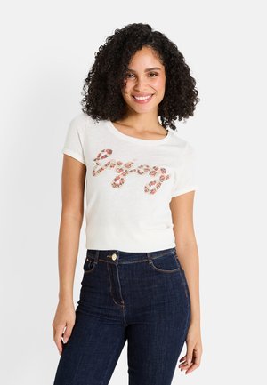 T-shirt blanc à manches courtes arborant un texte "Enjoy" dans un motif floral coloré. Accompagné d'un jean taille haute bleu foncé.