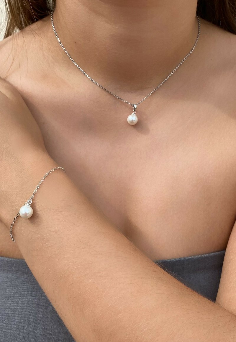 Silberne Kettenhalskette und passendes Armband mit einem einzelnen runden weißen Perlenanhänger. Glatte Textur und minimalistisches Design, auf der Haut getragen.