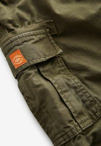 Olivgrön tygmanschett med sömmar och en vävd accent. Har en liten orange etikett med "Superdry"-logotyp.