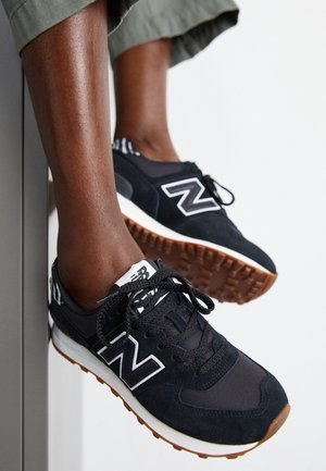 Osoba nosící černé tenisky New Balance s bílými a gumovými podrážkami, sedící se zvednutýma nohama na světlém pozadí.