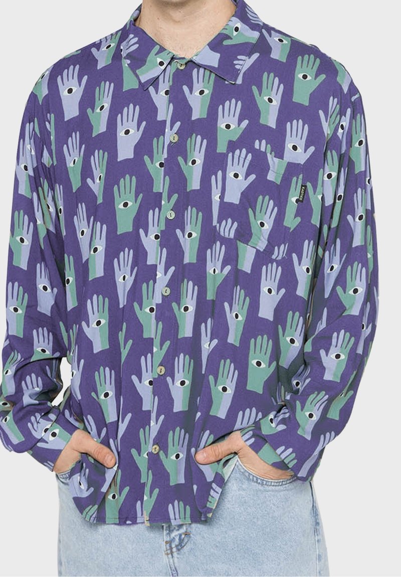 Homme portant une chemise violette à boutons avec des mains vertes et bleu clair présentant des yeux noirs dans les paumes, associée à un jean bleu clair.