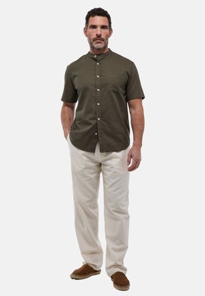 Chemise courte à manches courtes de couleur olive avec col mandarin, fermeture à boutons et poche poitrine unique. Pantalons crème et chaussures à enfiler marron.