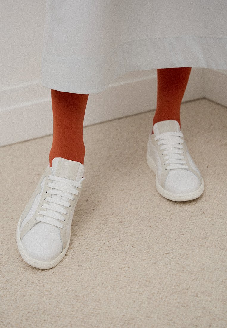 Stopy w białych trampkach i jasnopomarańczowych skarpetach pod białym ubraniem, stojące na beżowej wykładzinie w pobliżu narożnika białej ściany.