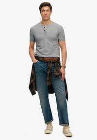 Szara koszulka henley z krótkim rękawem, niebieskie dżinsy, koszula w kratę zawiązana w talii, beżowe trampki za kostkę, brązowy skórzany pasek.