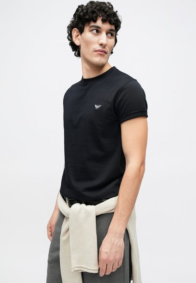 Juoda trumpomis rankovėmis marškinėliai su apvalia iškirpte, kairėje krūtinės pusėje yra mažas logotipas. Pilkos kelnės su šviesia megztine, rišama ties juosmeniu.