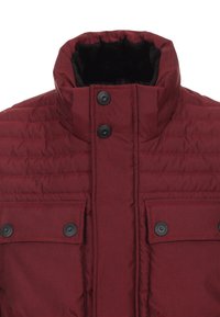 Burgundyröd puffjacka med hög krage, svarta knappar, ribbad textur och två framfickor med klaff. Fodrad med mörkt kontrasterande tyg.