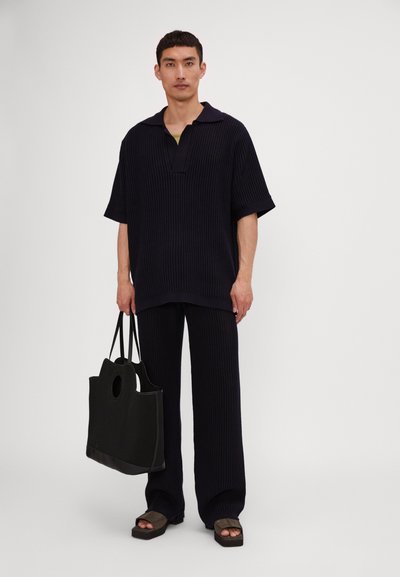 Polo shirt bleumarin tricotat cu mânecă scurtă și pantaloni evazați asortați, purtate cu o geantă de umăr neagră cu mânere decupate și un design texturat.