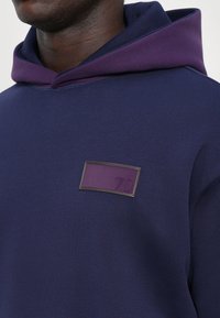 Námořnická modrá mikina s fialovou kapucí, se čtvercovou fialovou nášivkou s vytlačeným textem. Měkká textura a ležérní design.
