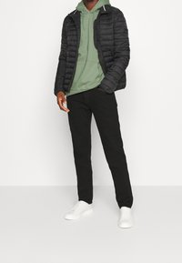 Veste matelassée noire portée sur un sweat à capuche vert clair, associée à un pantalon noir et des baskets blanches. La veste présente un design matelassé et une fermeture éclair à l'avant.