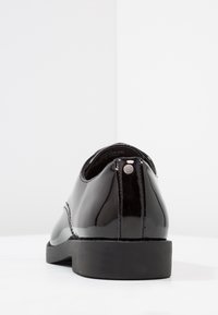 Chaussures en cuir verni noir avec une semelle plate, un bout arrondi et une couture arrière discrète. Elles présentent un petit accent argenté près du talon.