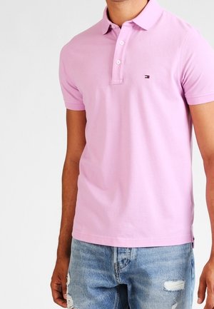 Muž v světle růžovém polo tričku s krátkým rukávem a malým logem na hrudi, doplněný o potrhané modré džíny.