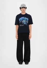 Tumesinine oversized T-särk, millel on sinine "VERSACE" graafika, koos mustade laiad püksid ja mustade kingadega, kantud heleda halli mütsiga.